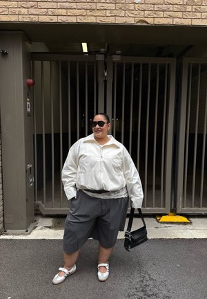 Persona con gafas de sol, chaqueta blanca, pantalones cortos grises y zapatos blancos, sosteniendo una bolsa negra, de pie frente a una puerta metálica cerrada.