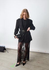 Blazer oversize nero con dettagli dorati, top nero, gonna in pizzo trasparente, tacchi neri e una piccola borsa nera. Sfondo semplice.