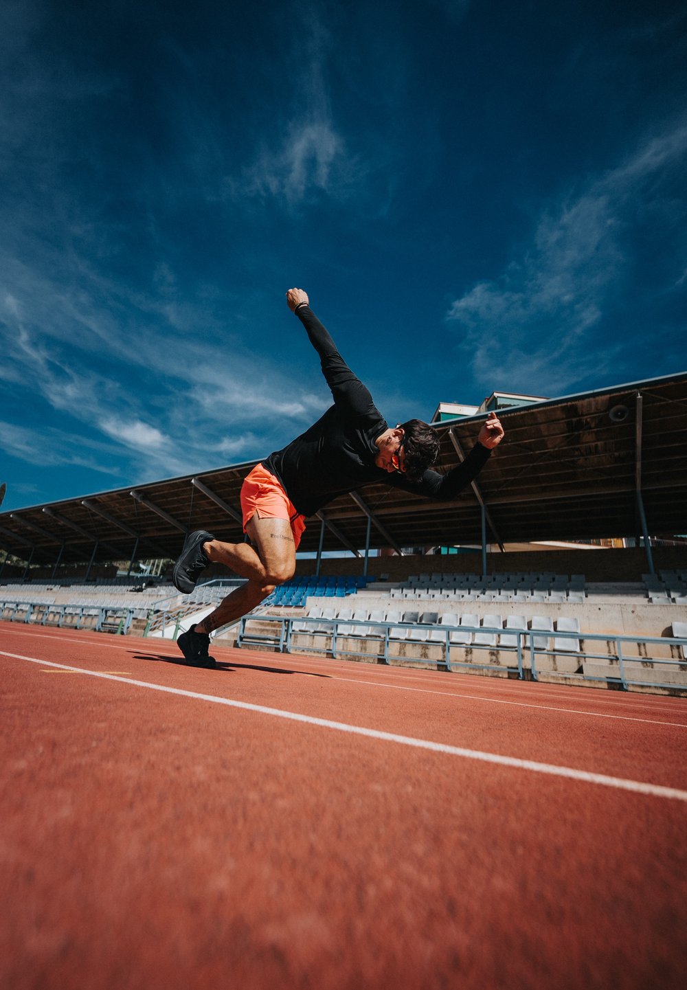 Läufer in schwarzem Oberteil und orangefarbenen Shorts, der von den Startblöcken auf einer Außenlaufbahn unter einem klaren blauen Himmel abspringt.