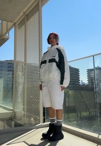 Giacca a vento bianca e verde, pantaloni corti bianchi, scarpe con tacco nero e calzini a righe, con uno sfondo urbano e un cielo azzurro limpido.