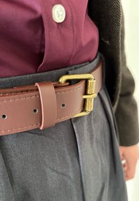 Curea din piele maro cu o cataramă din metal auriu, purtată peste pantaloni gri plisati și o cămașă de un burgund închis. Textură netedă cu cusături vizibile.