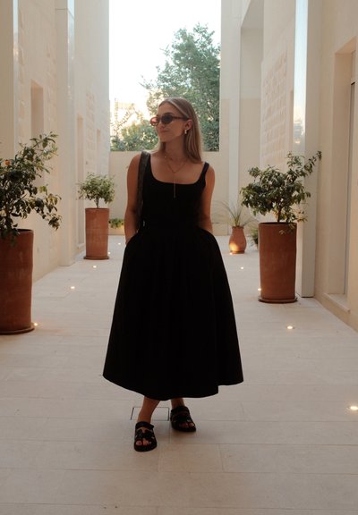 Vestido negro sin mangas con un corsé ajustado y una falda amplia, combinado con sandalias negras. El escenario presenta paredes de piedra pálida y plantas verdes.