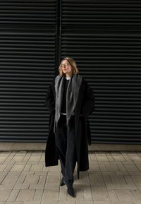 Long black coat over a grey sweater, paired with a grey scarf and black wide-leg trousers. Pointed black heels complete the outfit.