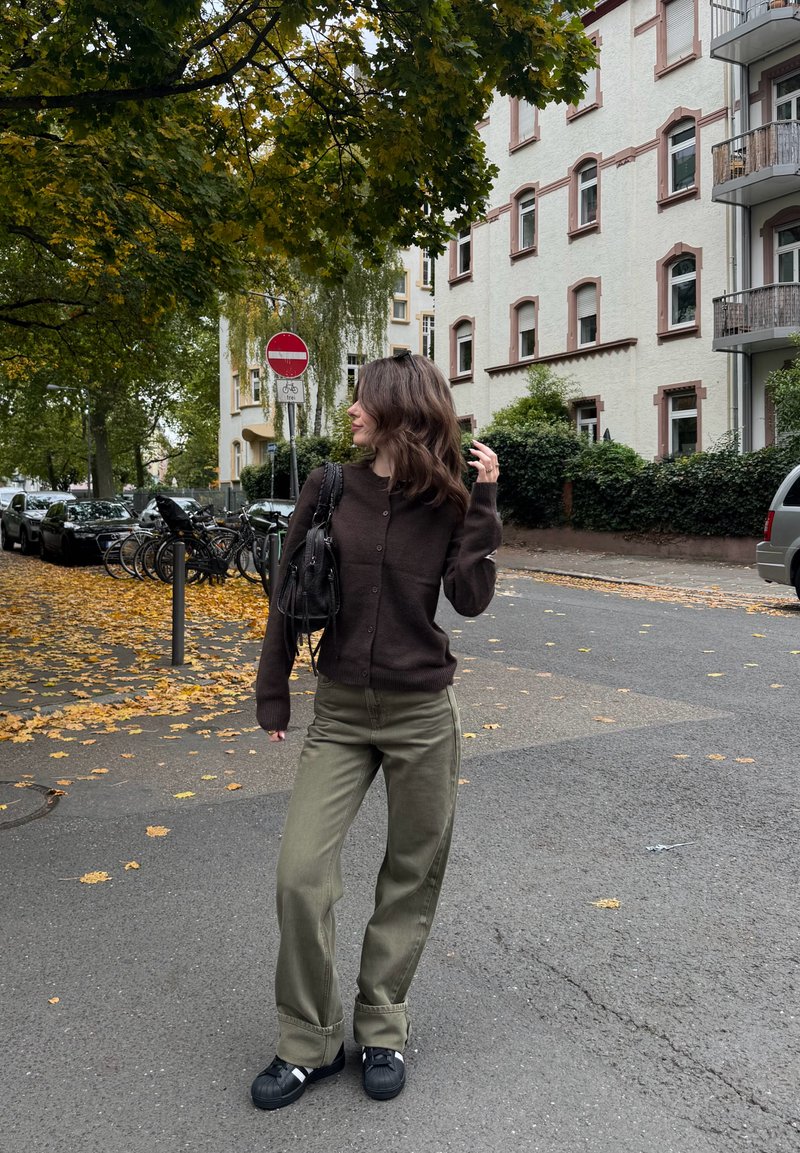 Cardigan marrone con bottoni, pantaloni verde oliva a gamba larga e scarpe da ginnastica nere. Foglie d'autunno e ambiente urbano con edifici e biciclette.