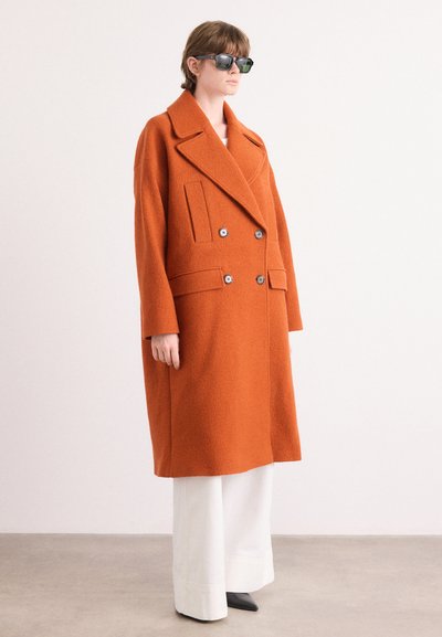 Abrigo de lana naranja con diseño de doble botonadura, grandes solapas, dos bolsillos delanteros y botones grandes. Combinado con pantalones de pierna ancha blancos y gafas de sol.