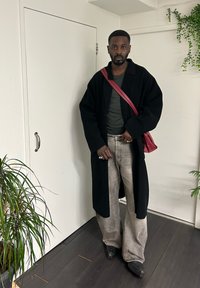 Άνδρας με μαύρο μακρύ παλτό, γκρι τζιν με πλαϊνές πιέτες, σκούρα παπούτσια και κόκκινη τσάντα ώμου στέκεται σε εσωτερικό χώρο κοντά σε φυτά και λευκούς τοίχους.