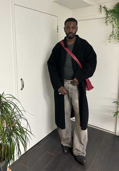 Hombre con abrigo negro largo, vaqueros grises de pierna ancha, zapatos oscuros y bolso de hombro rojo, de pie en el interior cerca de plantas y paredes blancas.