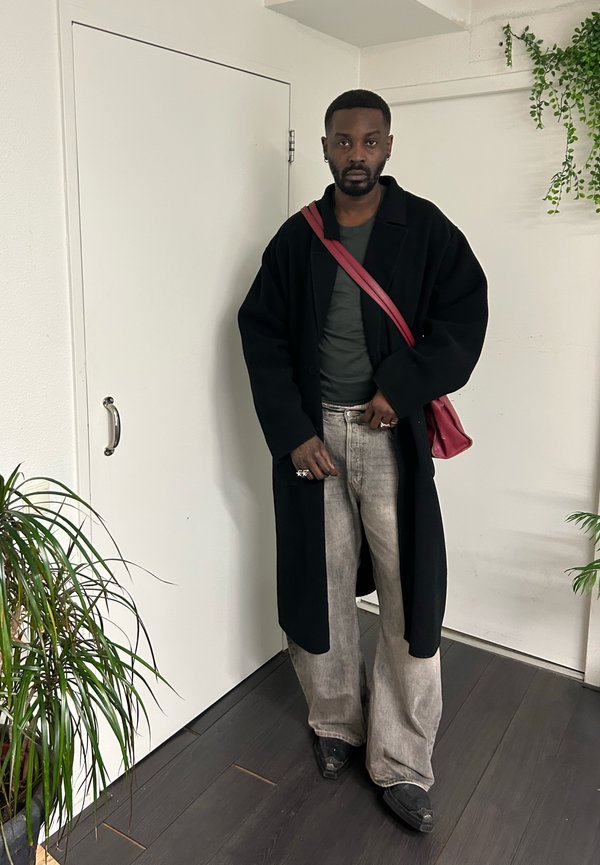 Hombre con abrigo negro largo, vaqueros grises de pierna ancha, zapatos oscuros y bolso de hombro rojo, de pie en el interior cerca de plantas y paredes blancas.