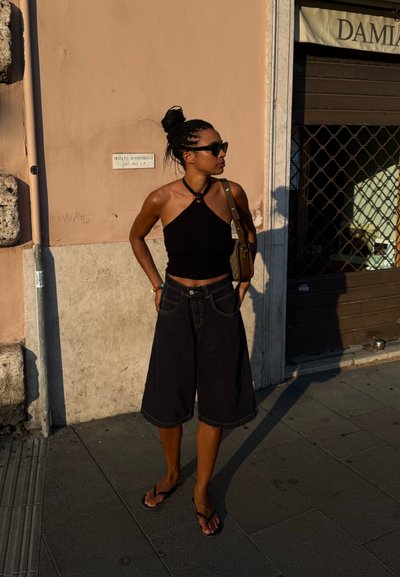 Top negro corto estilo halter combinado con pantalones cortos de denim anchos oscuros y chanclas. La figura está de pie frente a una pared rosa bajo la luz del sol.