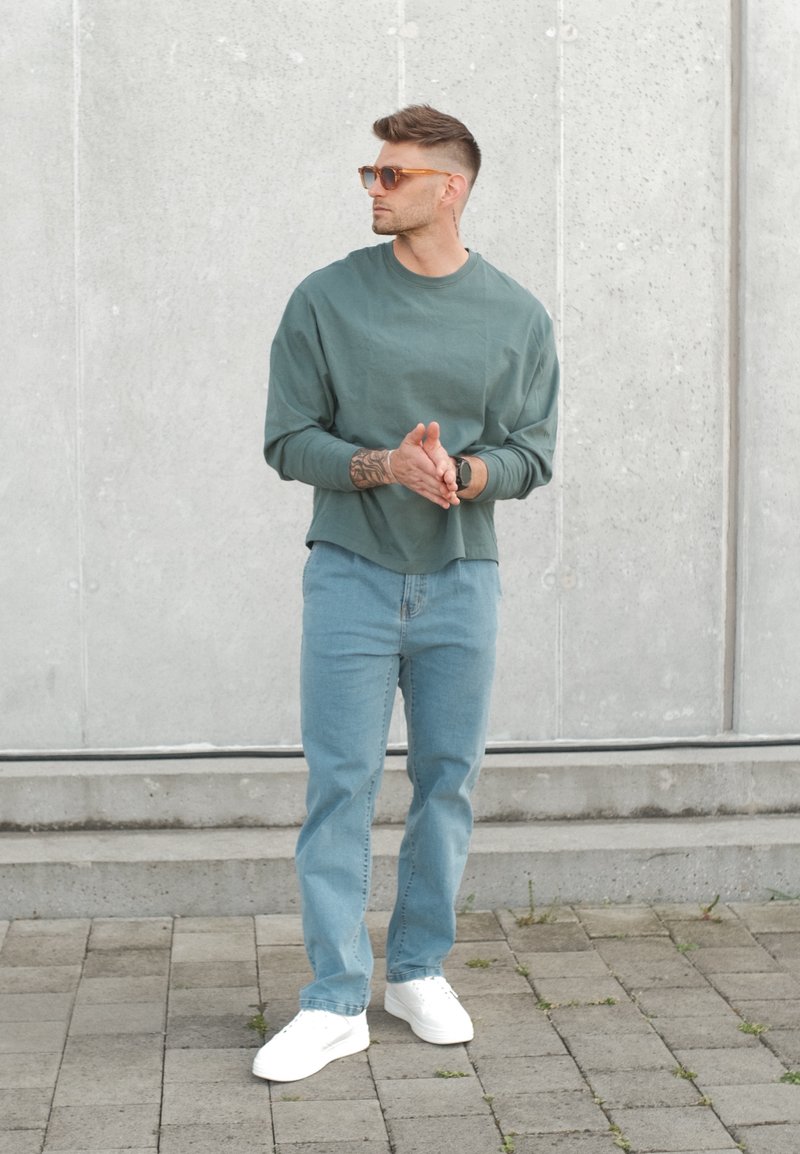 Homme portant des lunettes de soleil, une chemise verte à manches longues, un jean bleu et des baskets blanches, debout et regardant sur le côté contre un mur en béton.