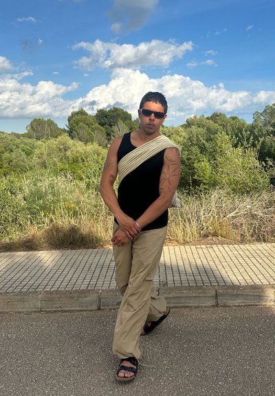 Top sin mangas negro, pantalones cargo beige y sandalias. Incluye un acento de hombro a rayas. Tatuajes visibles en el brazo izquierdo. Vegetación en el fondo.