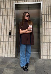Barna bordázott garbó poncsó, laza kék farmernadrág, fekete masszív cipő, kávéscsészét tartva, egy fém liftajtó mellett állva.