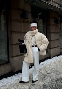 Femme portant un manteau et une écharpe en fausse fourrure couleur crème, pantalon blanc et béret, gants noirs et talons, tenant un sac à main noir dans une rue enneigée.