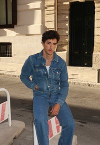Jaqueta de ganga e jeans em azul desbotado, com bolsos e costuras clássicas. Sentado sobre uma base de concreto perto de uma barreira de rua.