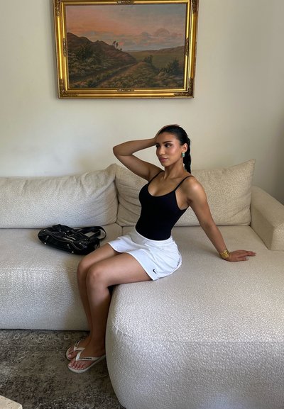 Mujer con camiseta de tirantes negra y falda blanca sentada en un sofá claro, apoyando una mano en la cabeza, con una pintura de paisaje en la pared encima.