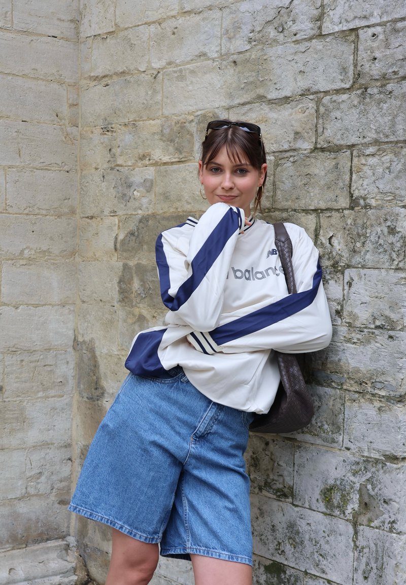 Giovane donna con occhiali da sole sulla testa, indossa una maglia bianca e blu a maniche lunghe e pantaloncini di jeans, appoggiata a un muro di pietra con una borsa marrone.