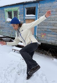 Veste en polaire crème chaude avec accents bleus, écharpe grise, jean noir évasé et chaussures foncées, debout sur la neige devant une cabane bleue.
