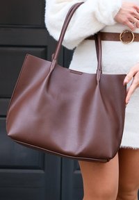 Femme portant une robe-pull crème avec une ceinture marron, tenant un grand sac fourre-tout en cuir marron à double poignée.