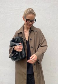 Brown oversized trench coat featuring large buttons, paired with a black textured bag and a black top. A plain white wall serves as the background.
