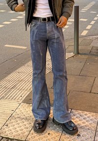 Personne portant un jean flare bleu, des chaussures noires à plateforme épaisse, une chemise blanche, une ceinture noire et une veste marron, debout sur un trottoir en ville.