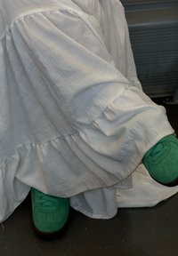 Sneakers in suede verde con design a lacci, suole nere a contrasto, abbinati a un abito bianco in tessuto testurizzato con orlo arricciato.