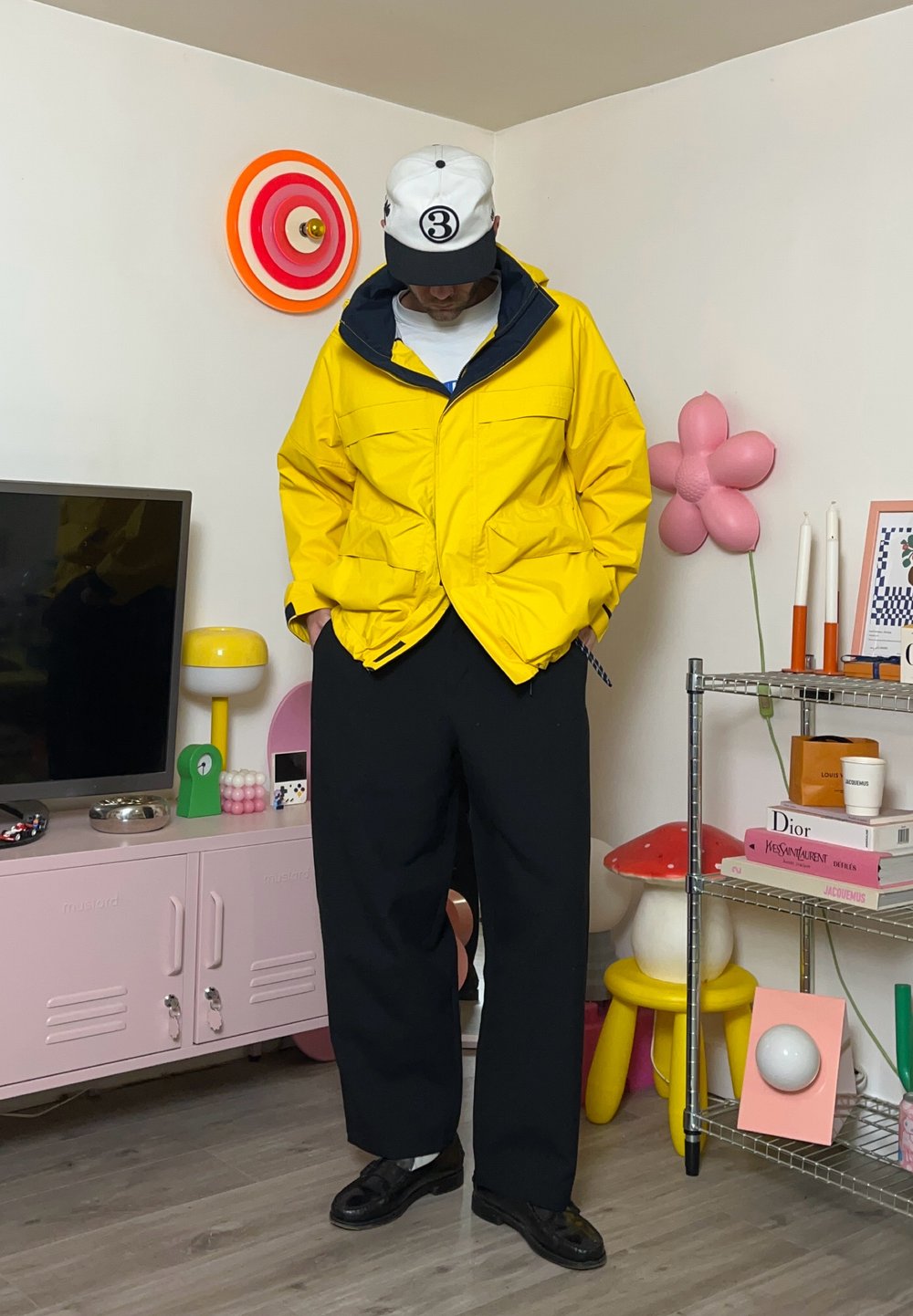 Giacca impermeabile gialla con colletto blu scuro, pantaloni neri a gamba larga e scarpe nere. Un cappellino bianco con logo nero completa l'outfit.