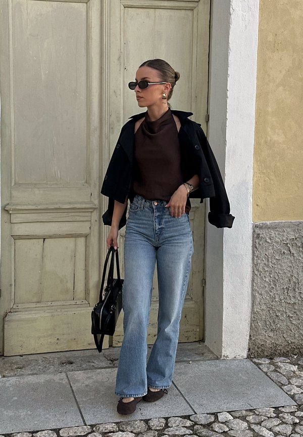 Kvinde iført solbriller, sort jakke over skuldrene, brun ærmeløs top, blå jeans, holder sort håndtaske, står foran en trædør.