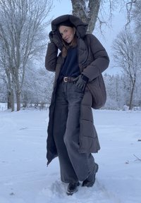 Femme portant un long manteau matelassé sombre, des gants, un jean large et des bottes, debout dans un paysage enneigé avec des arbres nus et un ciel crépusculaire.