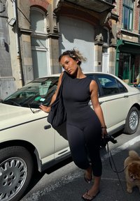 Mladá žena v čiernom bezrukávovom oblečení sa opiera o biely zaparkovaný automobil na mestkej ulici, drží vodítko pre psa s malým chlpáčom poblíž.