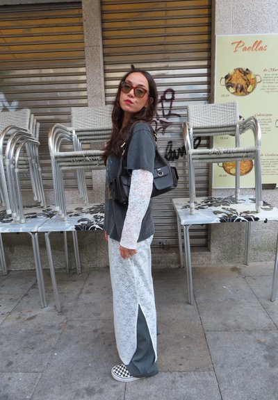 Camiseta gráfica gris, blusas largas de encaje blanco, pantalones a rayas gris y blanco con corte lateral, zapatillas de cuadros y un bolso negro de hombro.