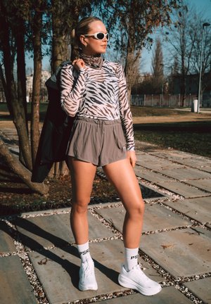 Mujer con camiseta de manga larga con estampado de cebra, pantalones cortos grises, zapatillas blancas y calcetines, de pie al aire libre sobre un camino pavimentado sosteniendo una chaqueta sobre el hombro.