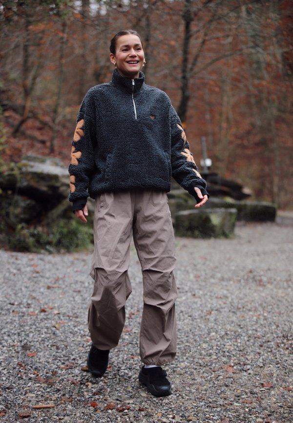 Woman smiling and walking on a gravel path in a forest, wearing a dark fleece jacket and light brown trousers in autumn.