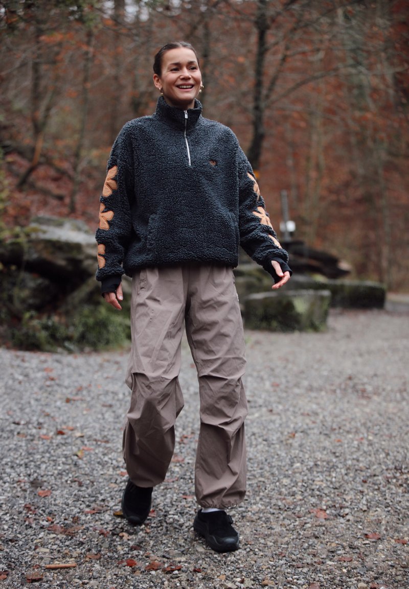Femme souriante marchant sur un sentier de gravier dans une forêt, vêtue d'une veste polaire foncée et d'un pantalon marron clair en automne.