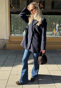 Jūros mėlynos spalvos dvigubas paltas, platėjantys džinsai, bateliai su leopardų raštu ir tekstūruota juoda rankinė, fone - saulėta gatvė.