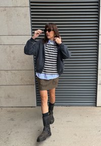 Chaqueta gris con capucha sobre una camisa de rayas, debajo una camisa azul con botones, falda corta oliva y botas negras hasta la rodilla con hebillas.