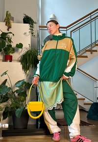 Πράσινο και μπεζ αδιάβροχο με λευκές λεπτομέρειες, συνδυασμένο με πράσινο παντελόνι. Κρατά μια κίτρινη τσάντα με σπειρωτή λαβή. Ροζ παπούτσια.