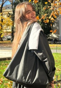 Chaqueta de cuero negra con detalles de forro polar gris, diseño extragrande, puños con botones y acabado elegante, usada sobre un vestido negro al aire libre.