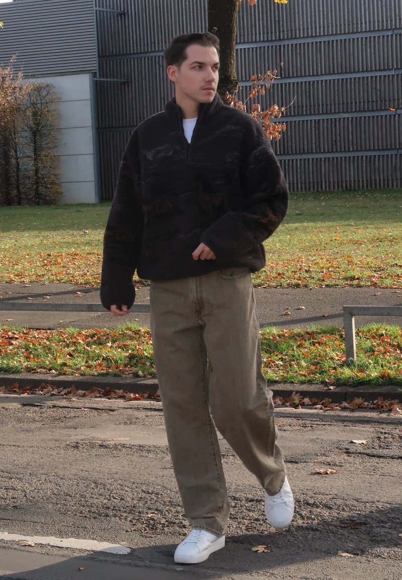 Polaire foncée à col montant avec un motif texturé, associée à un pantalon ample beige clair et des baskets blanches, marchant en extérieur.