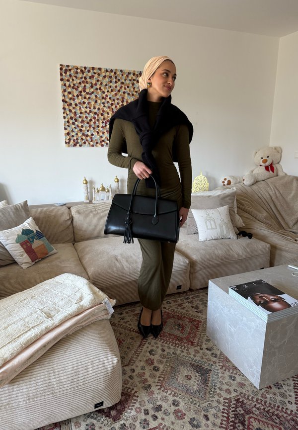 Mujer con vestido oliva y pañuelo en la cabeza está en la sala sosteniendo un bolso negro, con sofá beige, alfombra estampada y cojines decorativos.