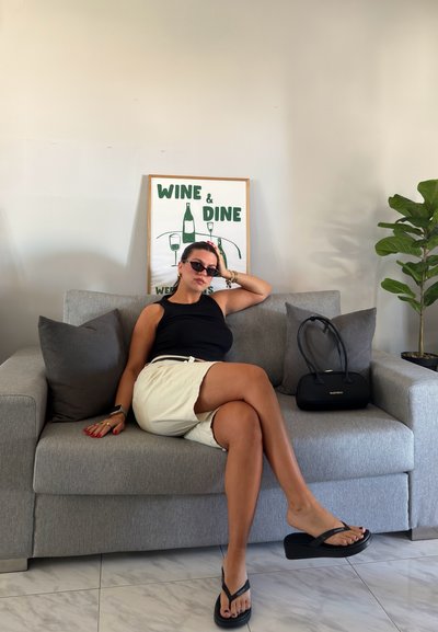 Mujer con gafas de sol y top negro sentada en un sofá gris con las piernas cruzadas, falda beige, bolso negro y cartel de "Wine & Dine" en la pared detrás.