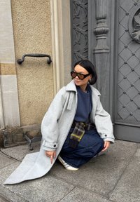 Szary wełniany płaszcz z dużym kołnierzem, niebieski sweter i ciemne dżinsy. Charakterystyczny żółto-czarny pasek w wzory; białe trampki w paski.