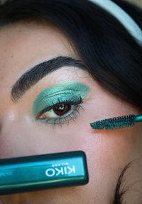 Teal mascara børste påfører produkt på øjenvipperne. Har grøn øjenskygge og brune øjne, med fine, veldefinerede vipper og en metallisk finish.