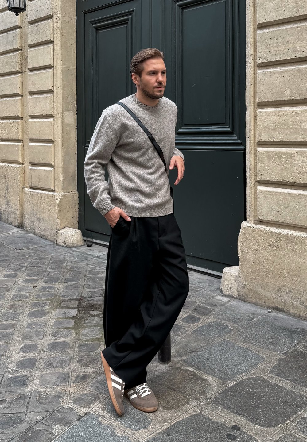 Grå strikket trøje, sorte brede bukser og brune sneakers med hvide striber. Står mod en struktureret væg, hænderne i lommerne.