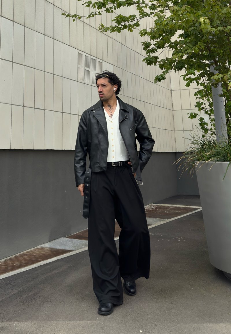 Mann trägt schwarze Lederjacke, weiße geknöpfte Weste, weite schwarze Hose und schwarze Schuhe und geht neben einem großen Pflanzkübel mit grünem Laub.