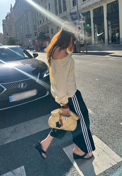 Conjunto informal que incluye un suéter suelto de color blanco roto, pantalones de chándal negros con franjas blancas y un bolso amarillo. Se llevan sandalias negras tipo slide.