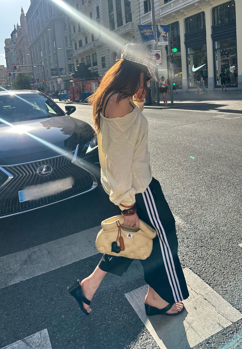Conjunto casual com um suéter solto em tom off-white, calças de treino pretas com riscas brancas e uma bolsa amarela. Sandálias pretas de slide. 