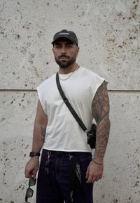 Weißes ärmelloses T-Shirt, dunkle Jeans und eine schwarze Umhängetasche. Accessoires umfassen eine Uhr, Sonnenbrille und ein schwarzes Bandana.