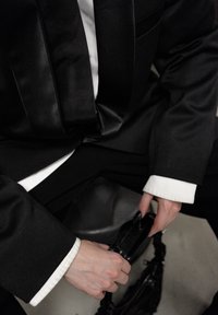 Smoking noir avec un nœud papillon en satin et une chemise blanche. Les mains tiennent un petit sac à main noir. L'arrière-plan est neutre et minimaliste.