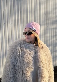 Femme portant un manteau beige duveteux, un bonnet rose avec une étiquette "GANNI" et des lunettes de soleil noires, debout contre un mur en métal ondulé.