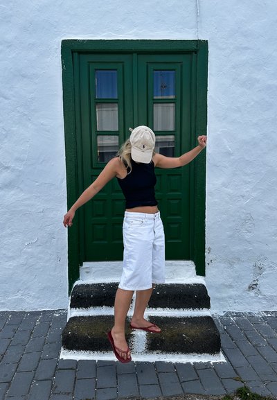 Pantalones cortos de denim blancos y una camiseta negra sin mangas, con una gorra de béisbol beige, de pie en unos escalones de piedra frente a una puerta verde y una pared blanca.